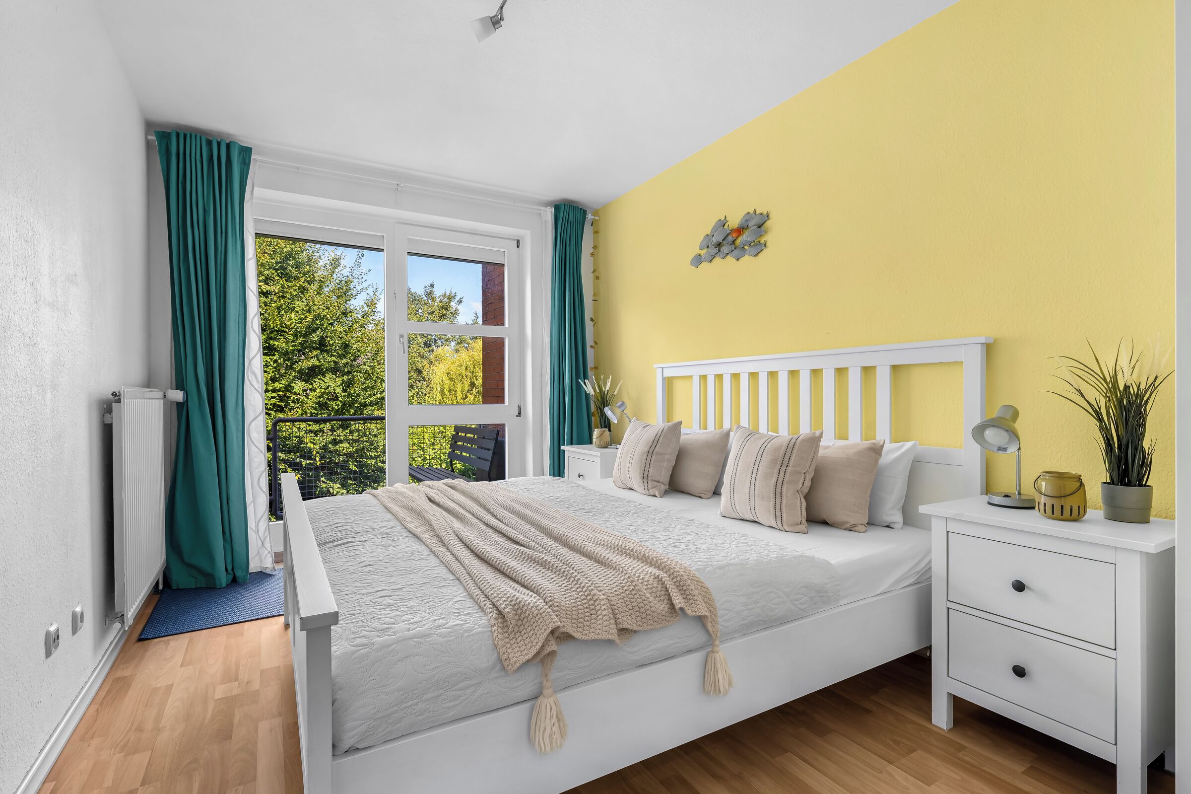 Un dormitorio con cama, mesita de noche, ventana con cortinas, radiador y vista a zonas verdes.