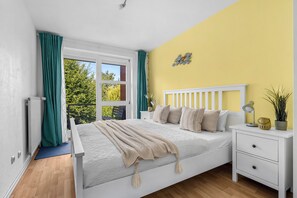 Ein Schlafzimmer mit Bett, Nachttisch, einem Fenster mit Vorhängen, einer Heizung und Blick auf Grünfläche.