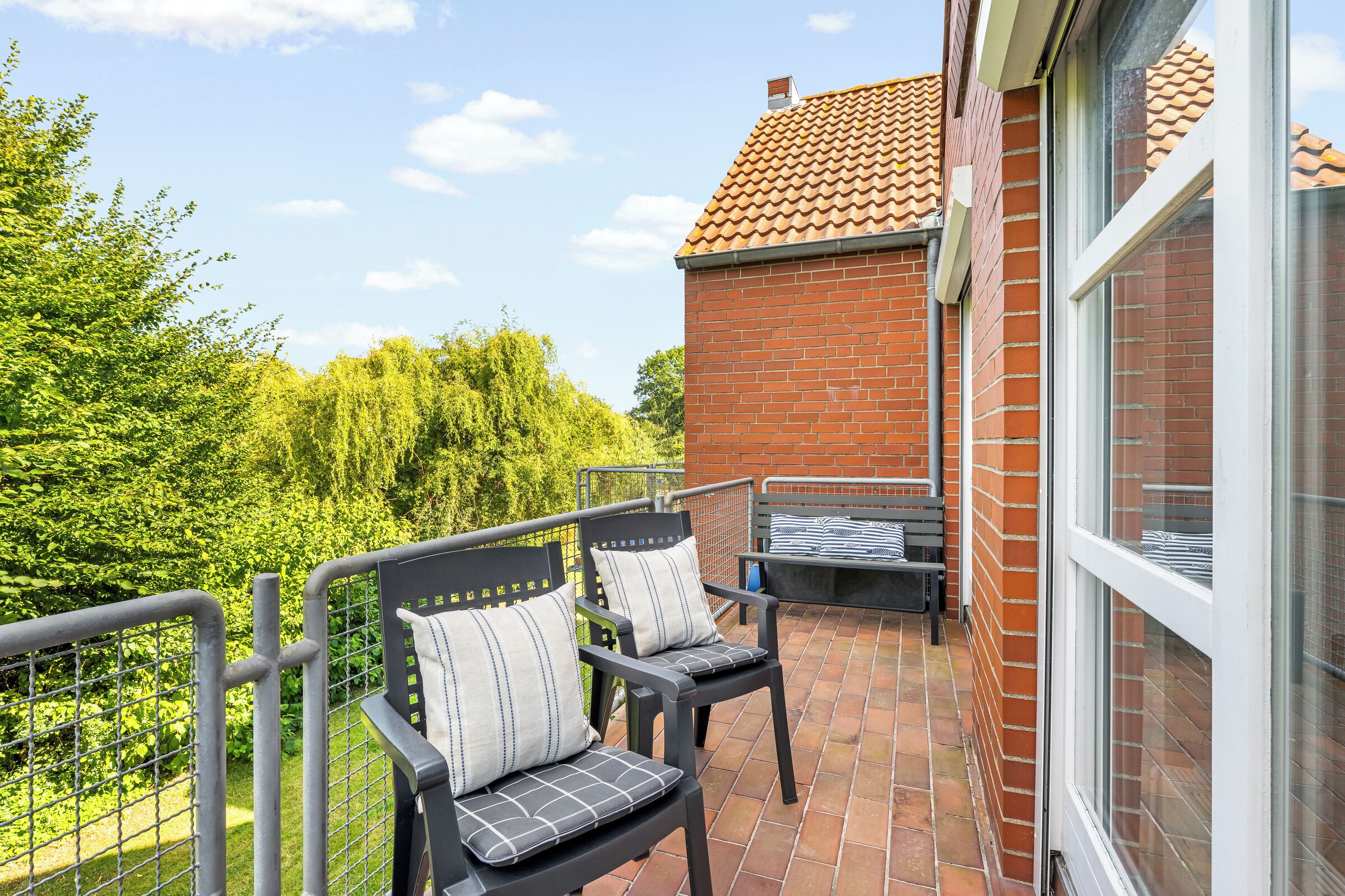 Ein Balkon mit zwei Stühlen und einem kleinen Tisch, mit Blick auf ein Backsteingebäude und Grünflächen.