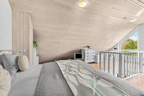 Ein Schlafzimmer mit schrägen Wänden, einem Bett mit gesteppter Matratze, einem Nachttisch, einer Kommode und einem Fenster mit Blick auf Grünflächen.