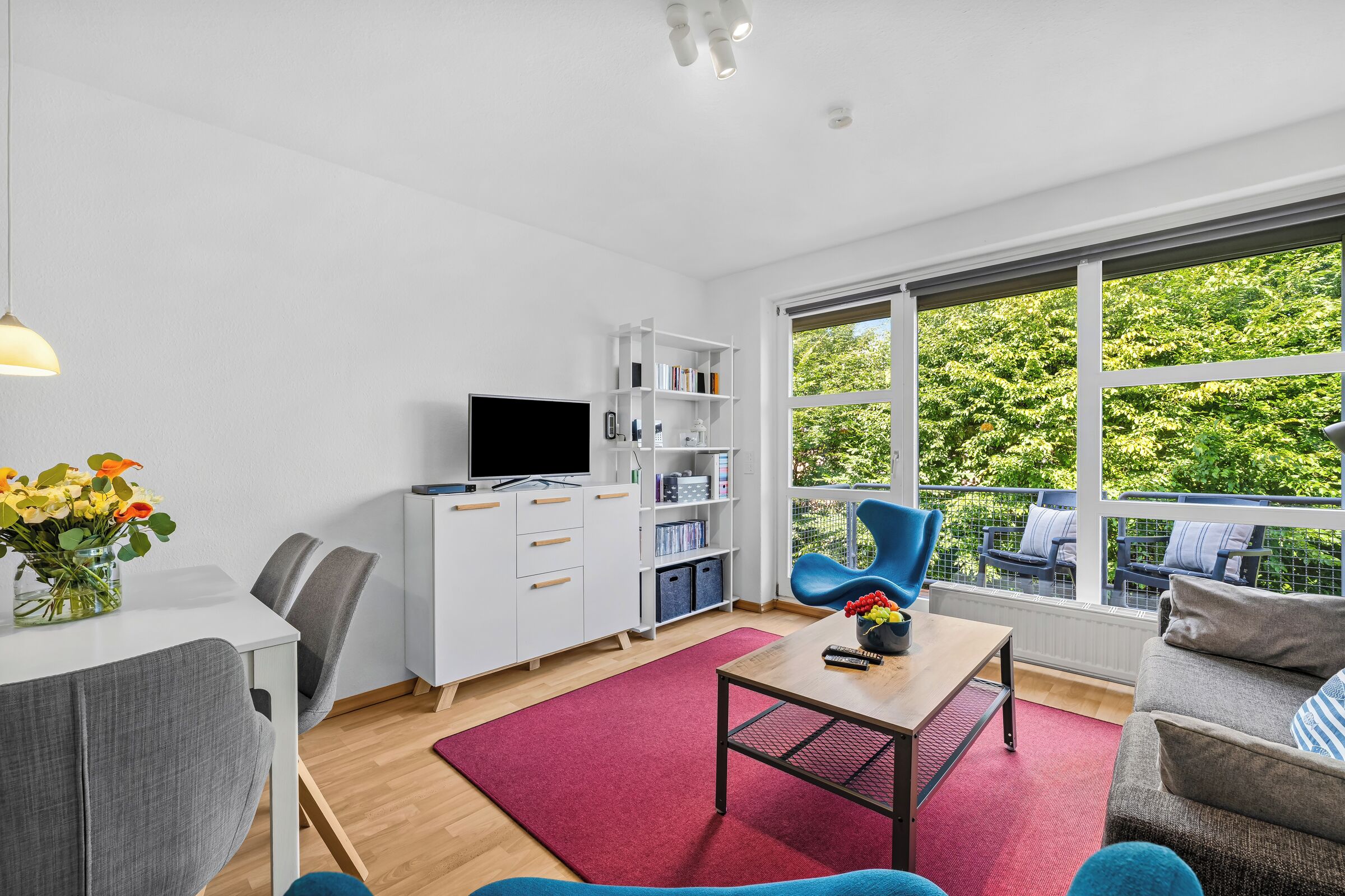 Ein modernes Wohnzimmer mit Flachbildfernseher, Bücherregal, Essbereich mit Tisch und Stühlen sowie ein großes Fenster mit Blick auf Grünflächen.