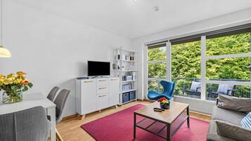 Ein modernes Wohnzimmer mit Flachbildfernseher, Bücherregal, Essbereich mit Tisch und Stühlen sowie ein großes Fenster mit Blick auf Grünflächen.