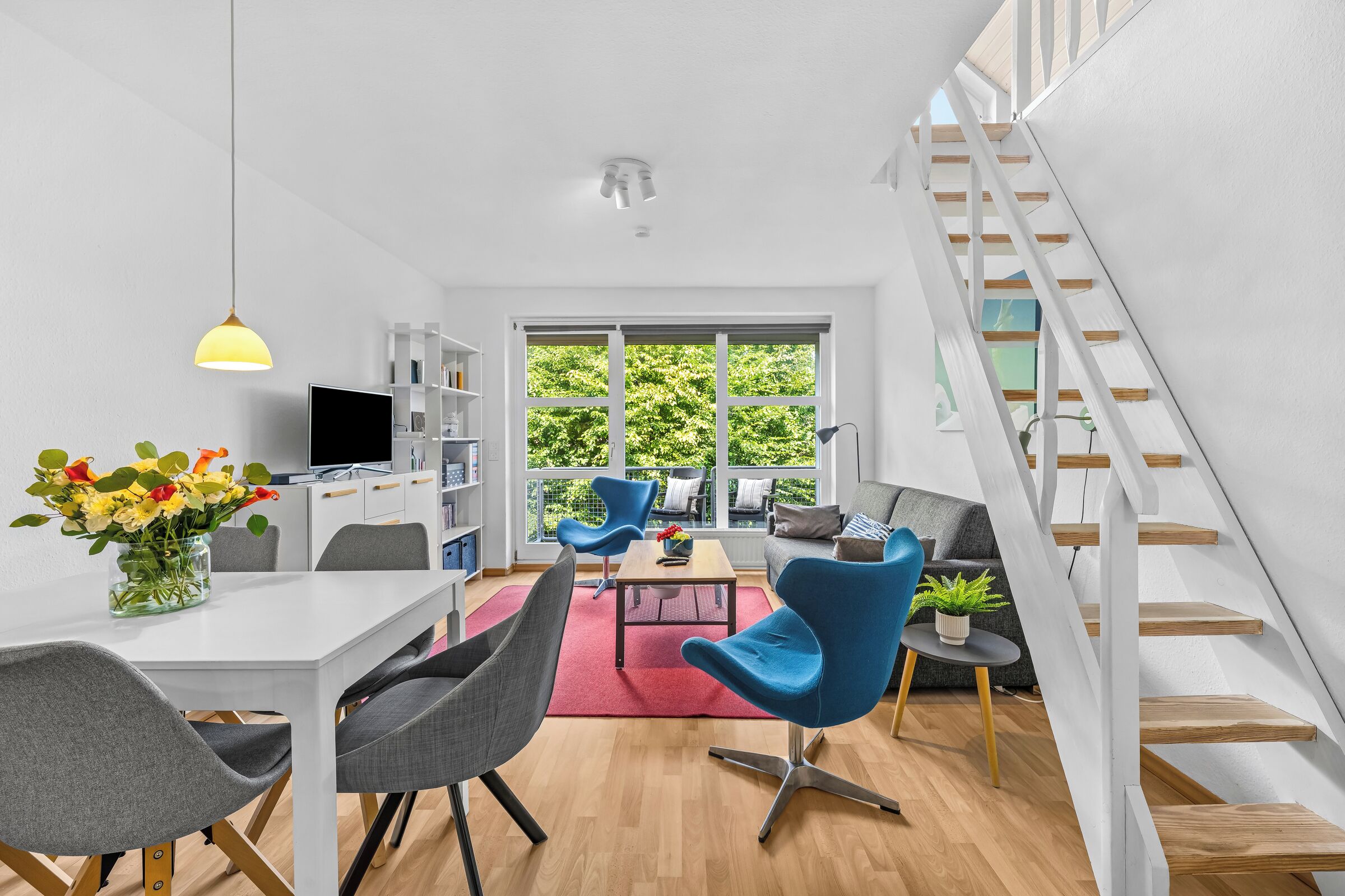 Ein modernes Wohnzimmer mit Essbereich, einer Treppe und einem großen Fenster mit Blick auf Grünflächen.