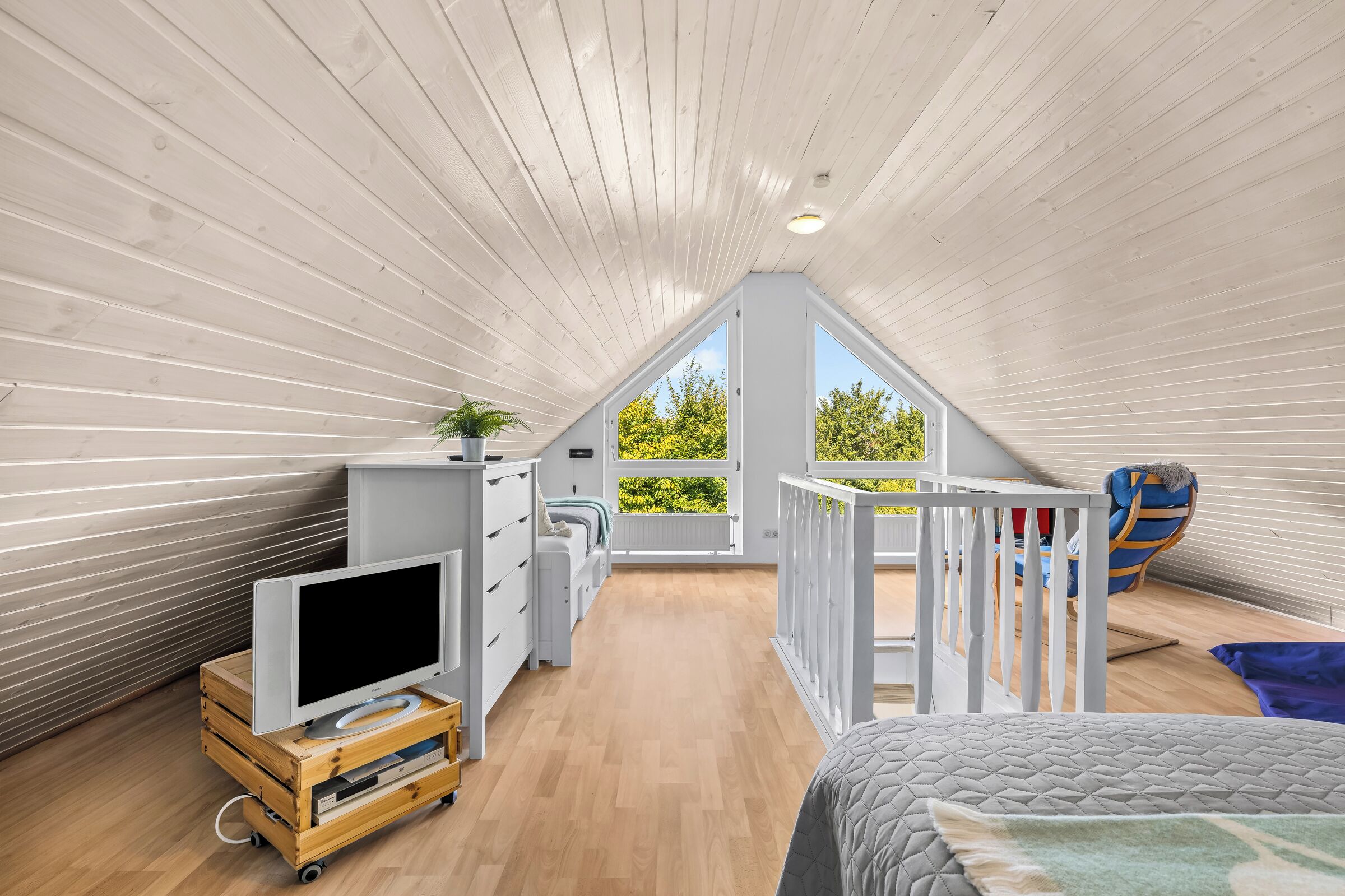 Une chambre spacieuse située dans les combles, avec un plafond en pente, une grande fenêtre donnant sur la verdure, un meuble TV en bois et un lit superposé.