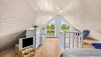 A spacious attic bedroom with a sloped ceiling, a large window offering a view of greenery, a wooden TV stand, and a bunk bed.