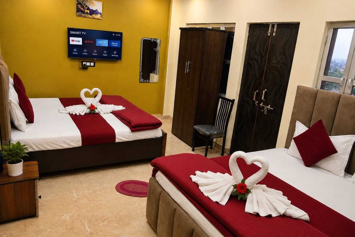 Habitación de hotel con dos camas, cada una decorada con una presentación de toallas con forma de cisne, un televisor mostrando servicios de streaming y vistas de la ciudad a través de una ventana.