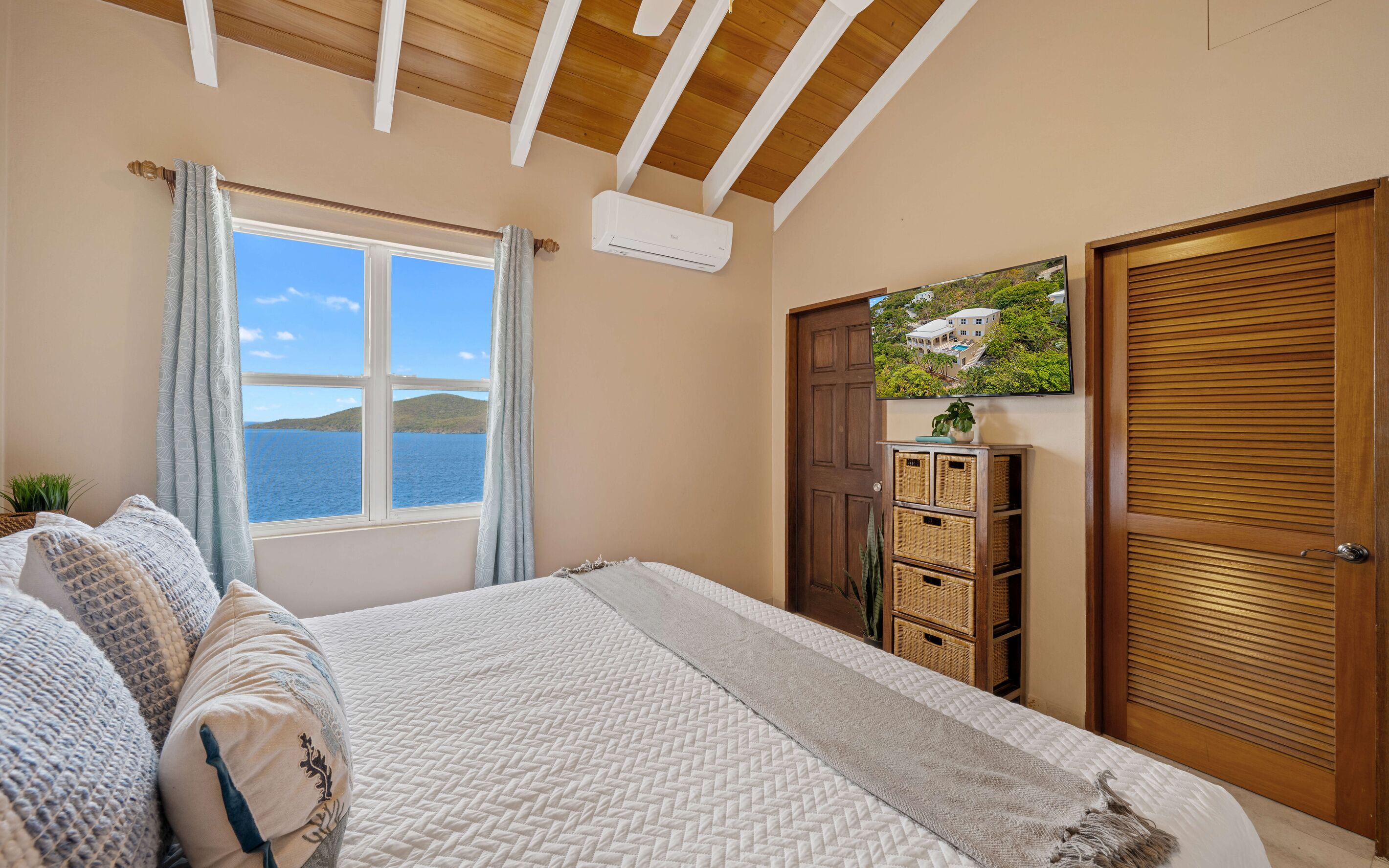 A bedroom with a large bed, a window with a view of the sea, a mounted television, and a wooden door.