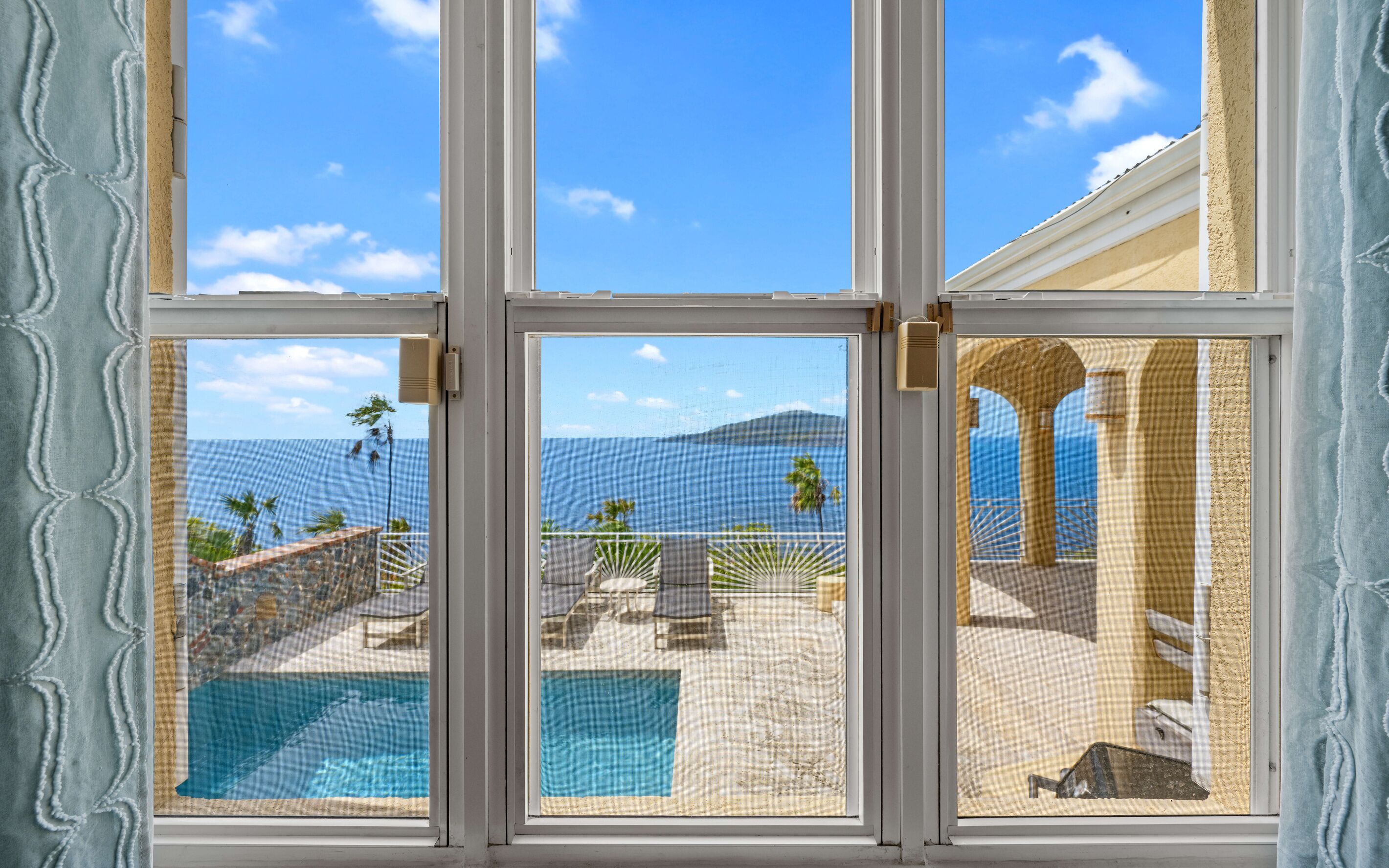A view through a window of a pool area with lounge chairs and a view of the ocean.
