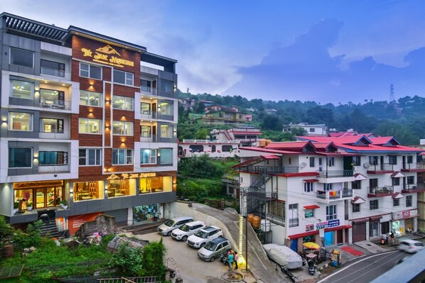 A multi-story hotel with a restaurant on the ground floor, parking in front, and a view of the surrounding hillside.