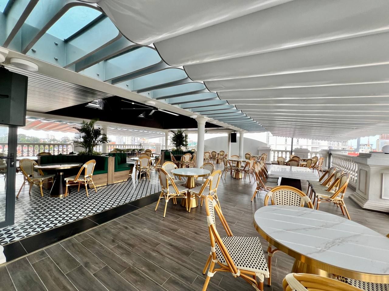 A spacious outdoor dining area with round tables and wicker chairs, a checkered floor pattern, and a modern architectural design with a glass ceiling.