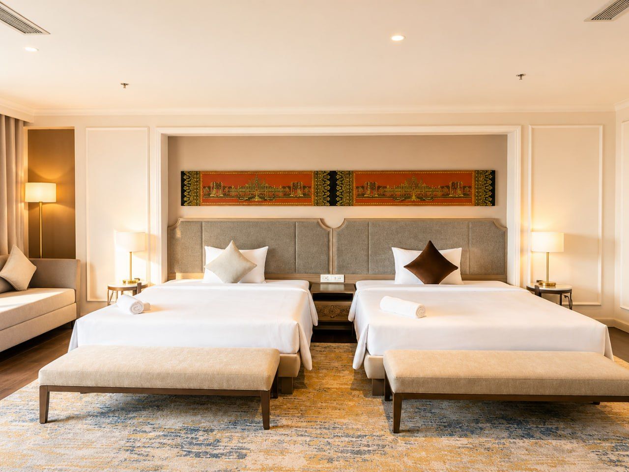 A hotel room with two beds, a sofa, a large painting on the wall, and a patterned rug.