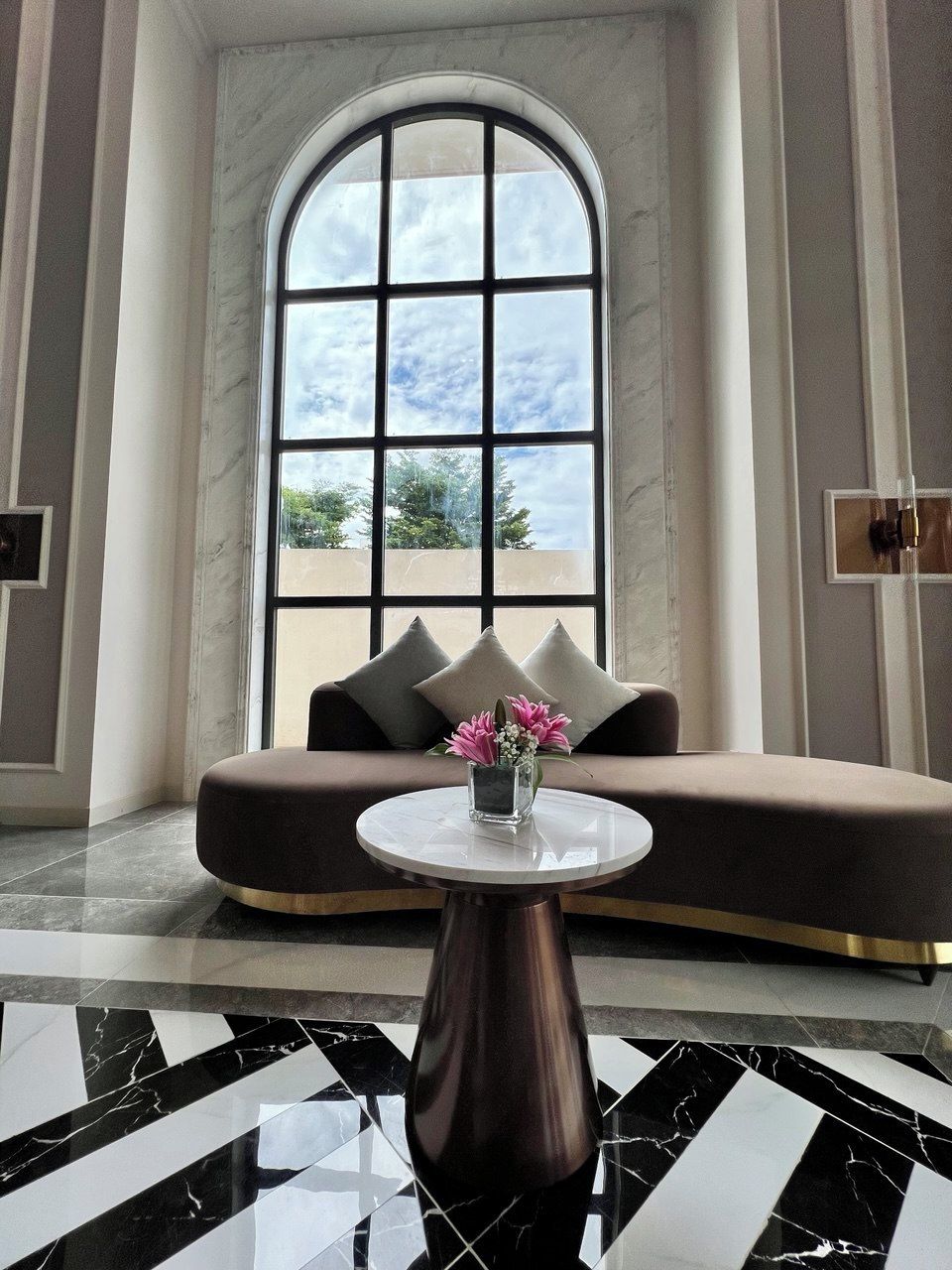 A modern lobby with a large arched window, a sofa with cushions, and a small table with a vase of flowers.