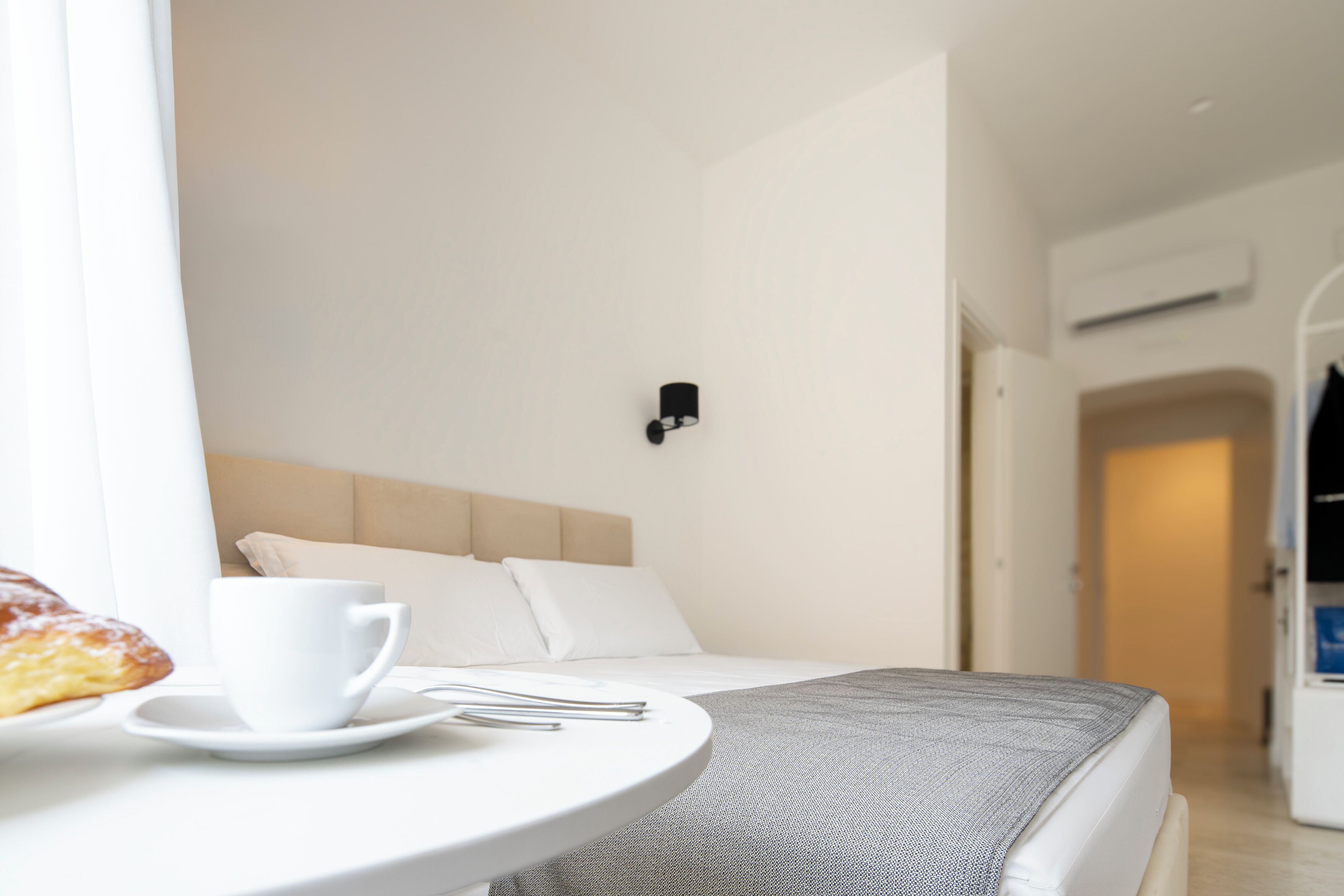 Habitación de hotel con cama, taza y platillo blancos, y una mesita con plato y cubiertos.