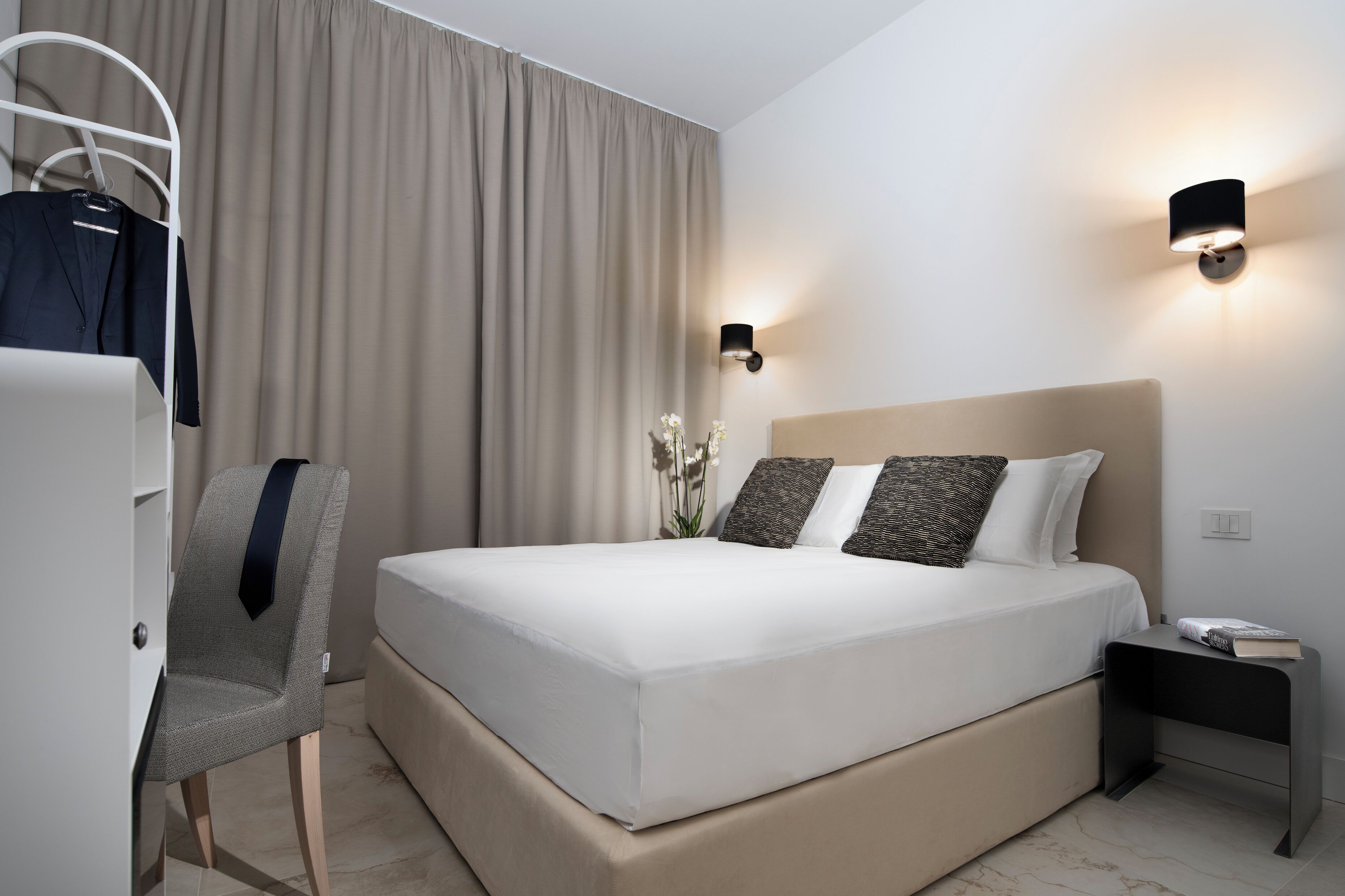 A modern hotel room with a large bed, a bedside table, a chair, and a wardrobe.