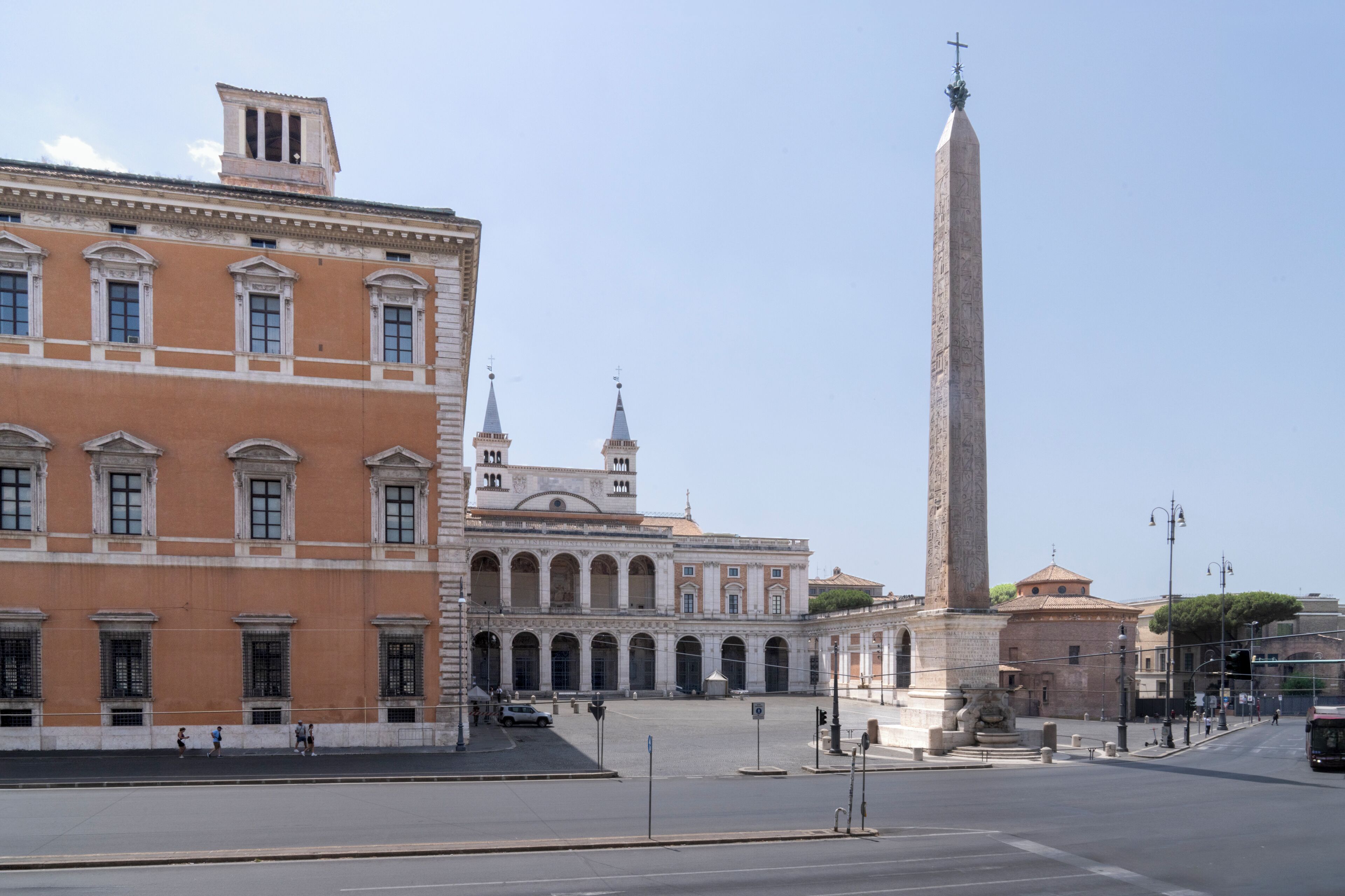 Un bâtiment historique, avec un obélisque imposant devant, un ciel dégagé et une rue où l’on aperçoit quelques piétons.