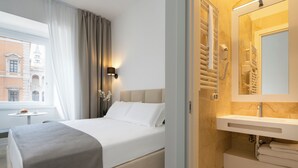 A hotel room with a bed, a bedside table, a lamp, a window with curtains, and a bathroom with a sink and mirror.