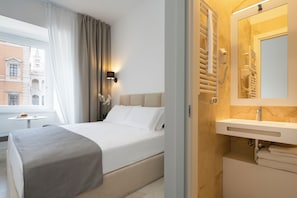 Habitación de hotel con cama, mesita de noche, lámpara, ventana con cortinas y baño con lavabo y espejo.