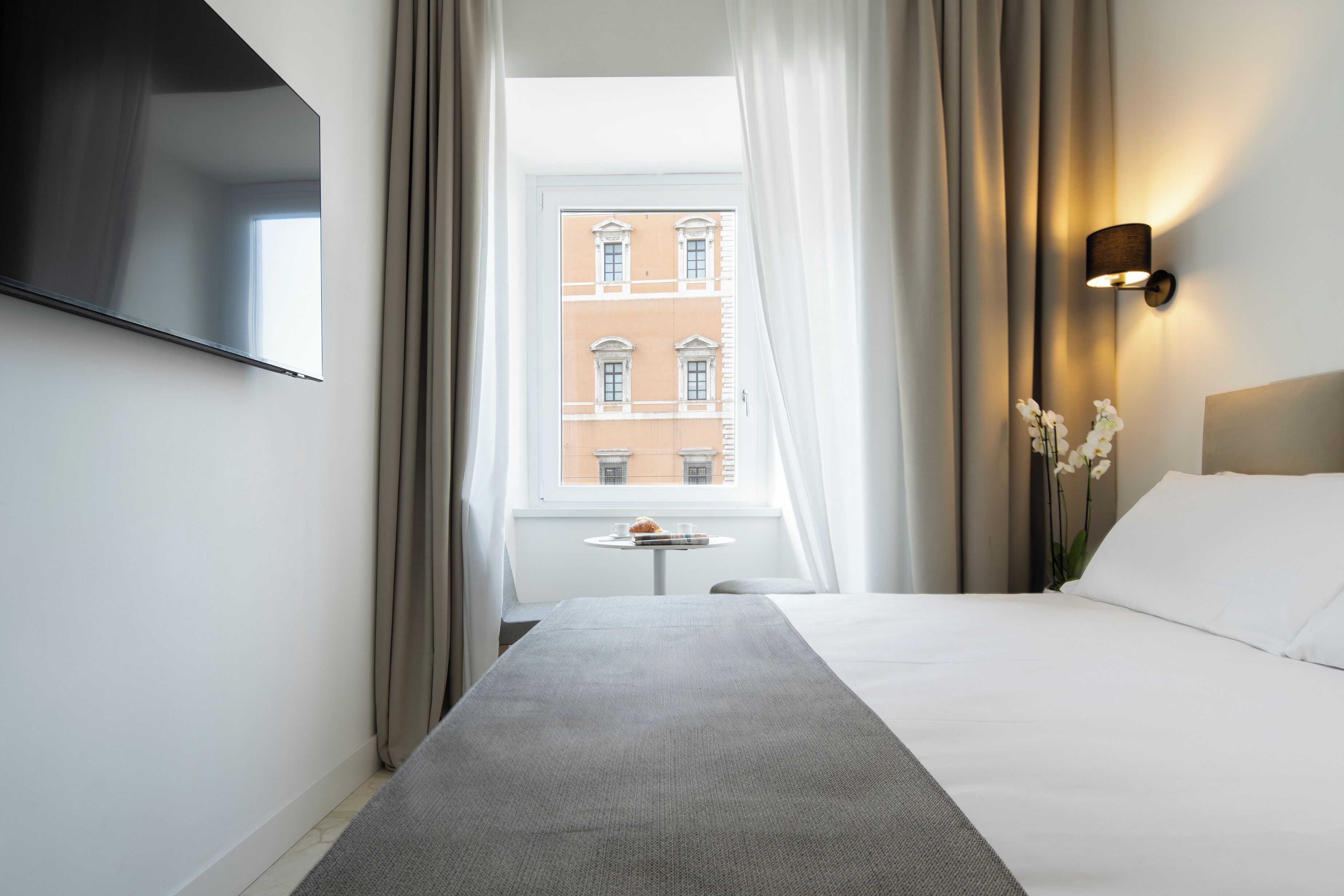 Une chambre d’hôtel avec un lit, un téléviseur fixé au mur, une petite table avec une tasse et un livre, et une fenêtre avec des rideaux.