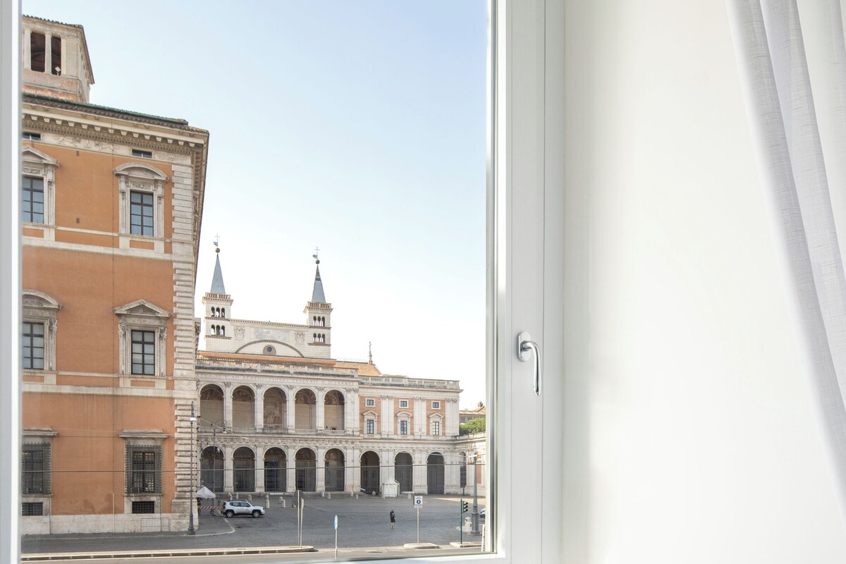 Una habitación con ventana que da a un edificio histórico, una pequeña mesa redonda con alimentos para el desayuno y un periódico.