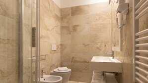 A bathroom with a glass shower enclosure, a bidet, a sink with a towel rack, and a towel dispenser.