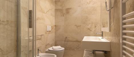 A bathroom with a glass shower enclosure, a bidet, a sink with a towel rack, and a towel dispenser.