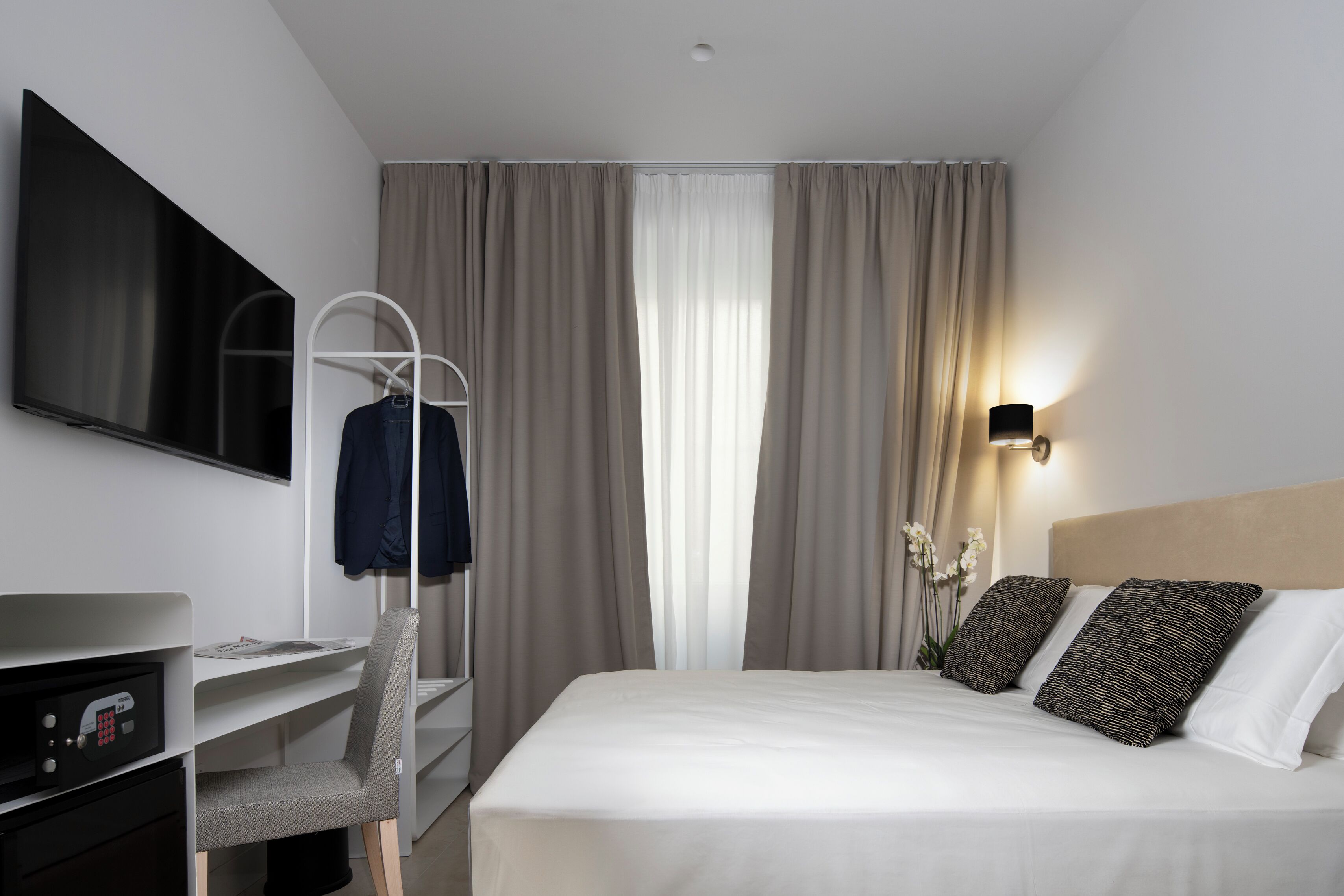 A hotel room with a bed, a desk with a chair, a TV mounted on the wall, and a window with curtains.