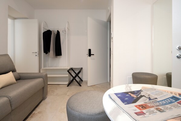 A living room with a sofa, a coffee table with newspapers, a small table with a tray, and a door leading to a bathroom.