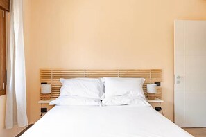 Ein ordentlich bezogenes Bett mit weißen Leinen, einem Holzkopfstück und je einem Nachttisch mit Lampe auf beiden Seiten.