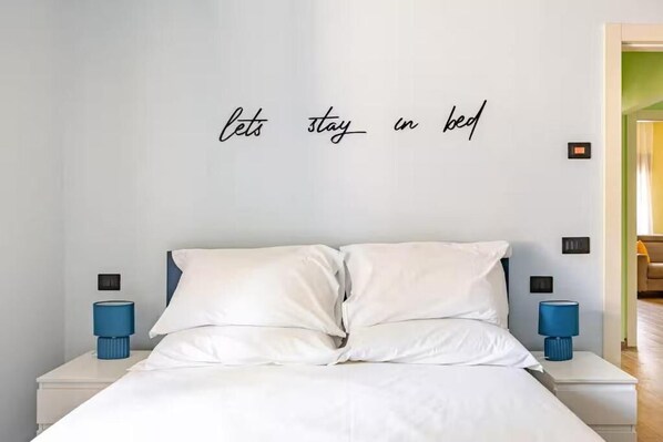 A bed with white bedding and two blue bedside lamps, a wall with a sign that says 'lets stay in bed', and a door leading to another room.