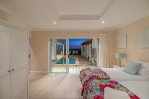 A hotel room with a bed, a nightstand, a lamp, and a view of a pool and sunset through a glass door. - Pezula Fairlead Luxury 7A  (Knysna)