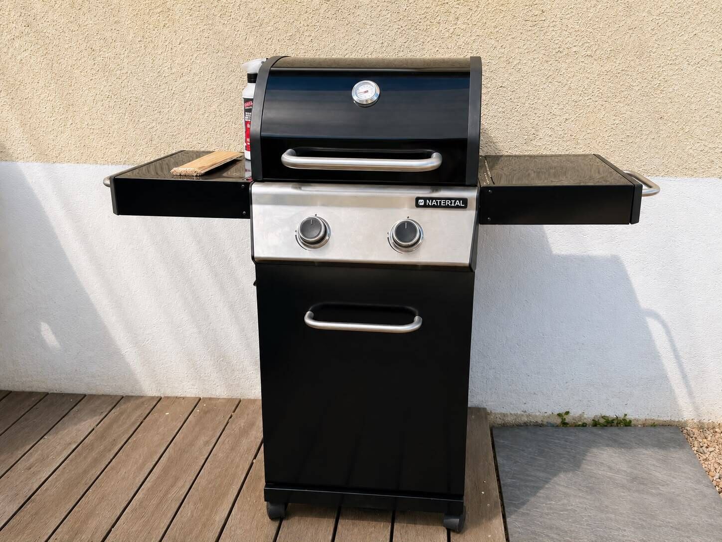 A black barbecue grill with a digital thermometer and two control knobs, placed on a wooden deck against a white wall.