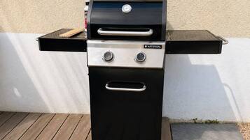 A black barbecue grill with a digital thermometer and two control knobs, placed on a wooden deck against a white wall.