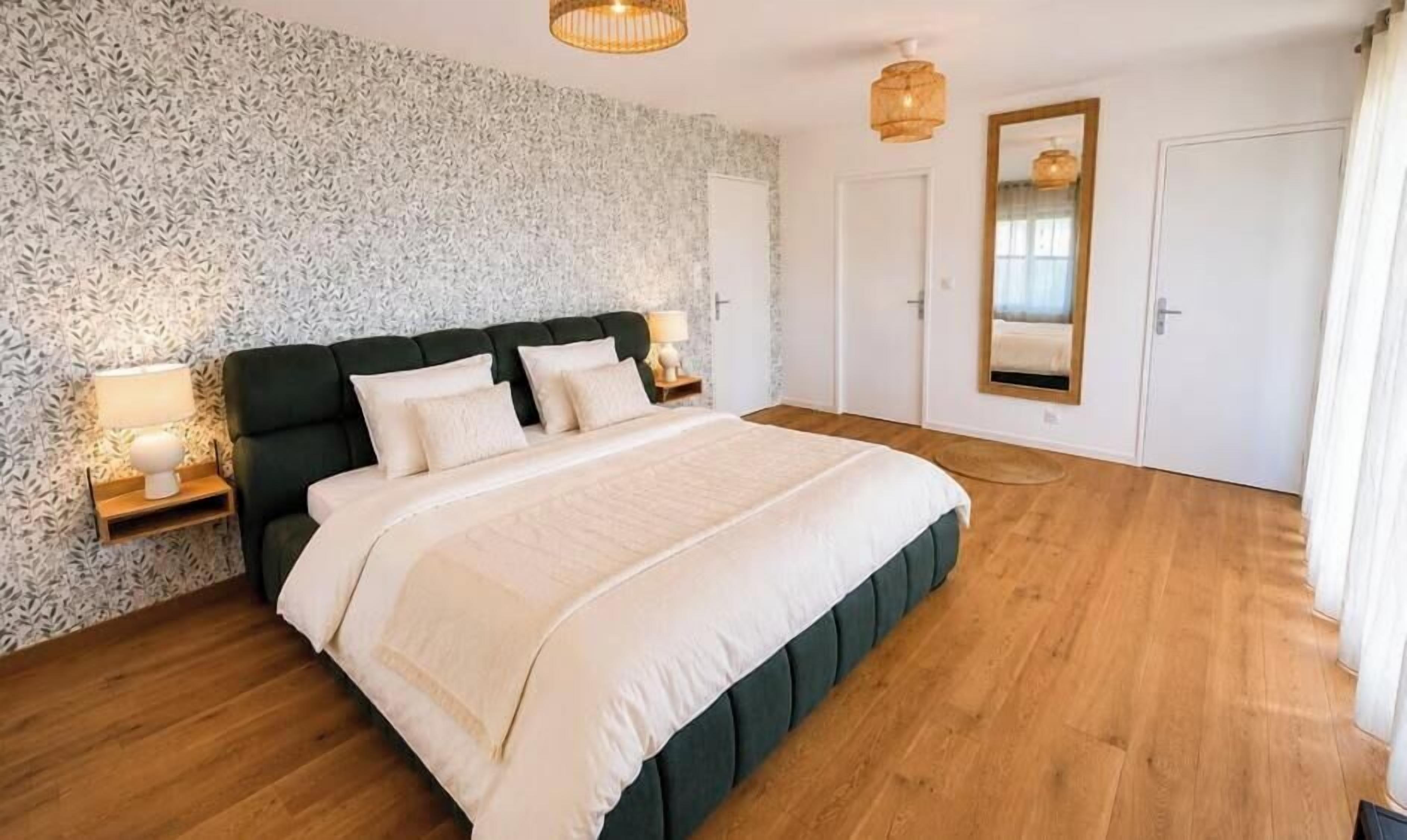 A bedroom with a large bed, a nightstand, a wall-mounted mirror, and a door.