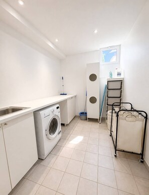 A laundry room with a washing machine, a sink, a drying rack, and storage bins.