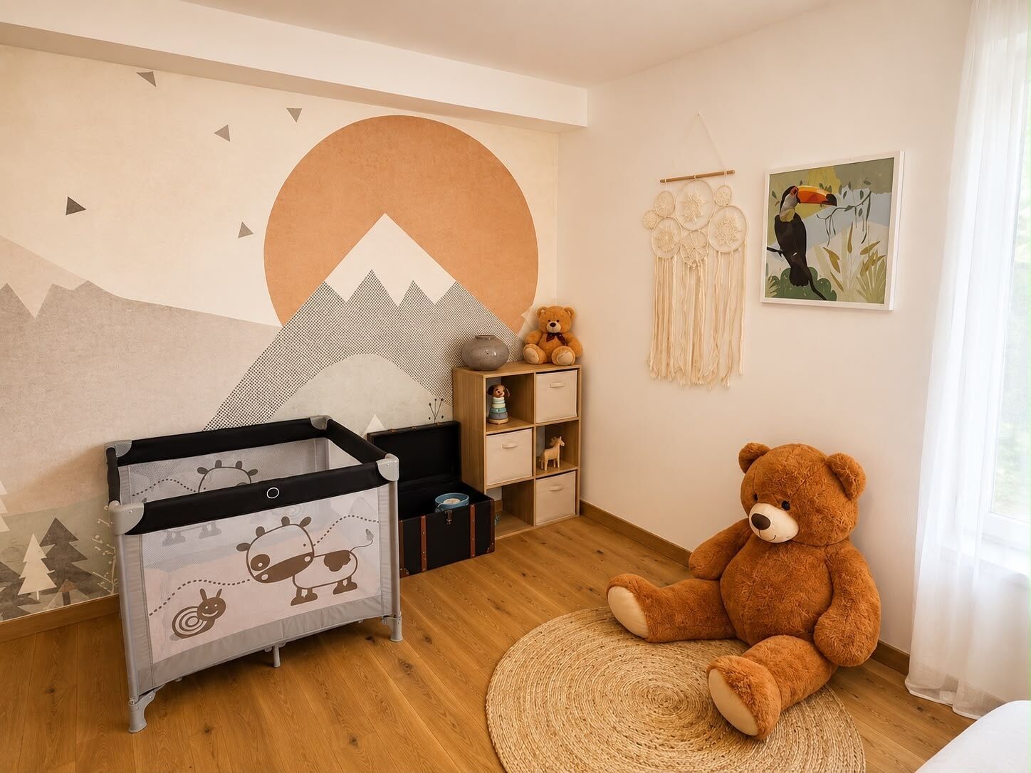 A child's room with a crib, a large teddy bear, a dreamcatcher, and a mountain mural on the wall.