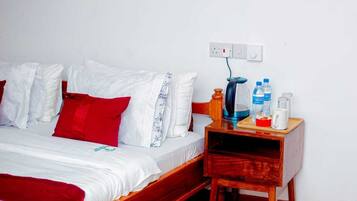 A single bed with white and red bedding, a wooden nightstand with a kettle, water bottles, and a mug.
