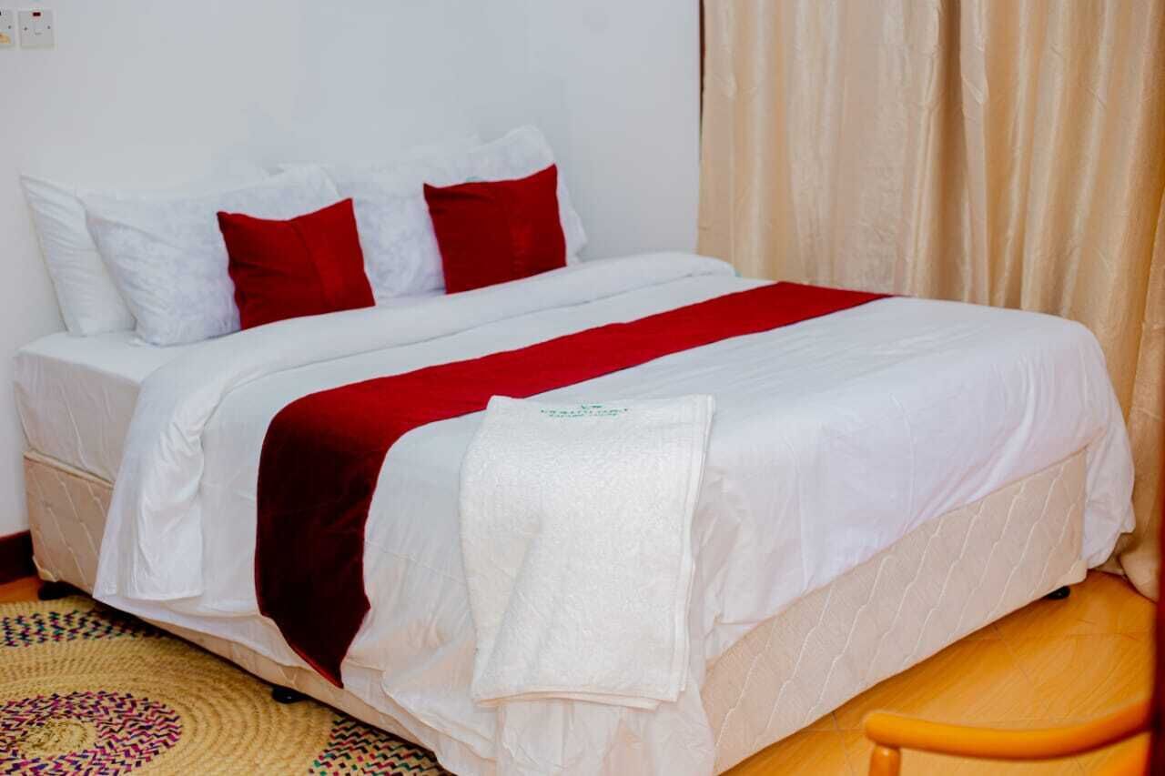 A bed with white bedding and red accents, a white towel, and a woven floor mat.