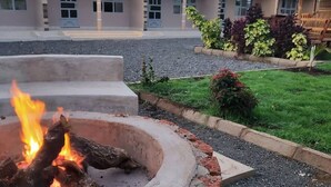 A fire pit with burning logs, surrounded by a concrete border, in front of a building with a thatched roof and a paved pathway.