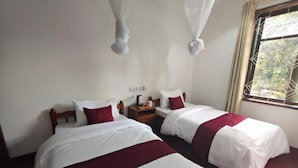 A hotel room with two beds, a nightstand, and a window with a view of greenery.