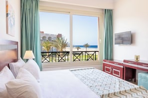 Ein Hotelzimmer mit Bett, Schreibtisch, Fernseher und einem Balkon mit Blick auf das Meer und Gebäude.