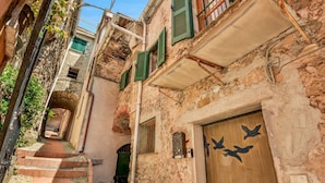 Ein schmaler Gang mit einer Holztür, die ein Vogelmotiv zeigt, begrenzt von Gebäuden mit Steinmauern und grünen Fensterläden.