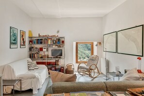 Ein modernes Wohnzimmer mit einer Couch, einem Bücherregal, einem Fernseher und einem Fenster mit Blick auf Bäume.