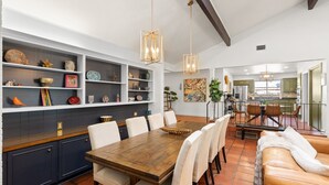 A spacious dining area with a wooden table and chairs, a kitchen in the background, and decorative shelves with various items.