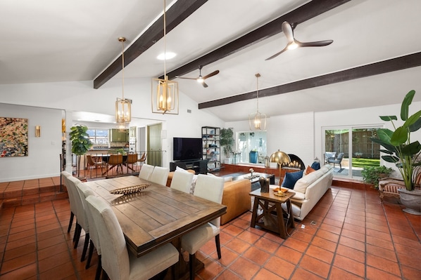 A spacious living area with a dining table, sofas, and a TV. There are ceiling fans and pendant lights.