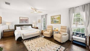 A bedroom with a bed, two armchairs, a ceiling fan, and a window with curtains.