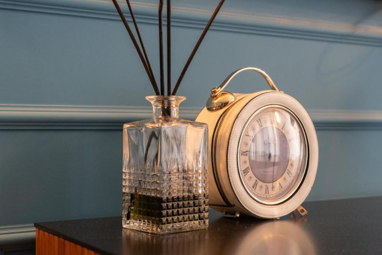 Una botella de vidrio transparente con superficie texturizada y un reloj despertador metálico redondo con números romanos en la esfera, sobre una superficie reflectante.