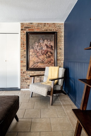 A room with a brick wall, a large painting, a patterned armchair, and a wooden bench. - The Getaway - Sleeps 12 (Belton)
