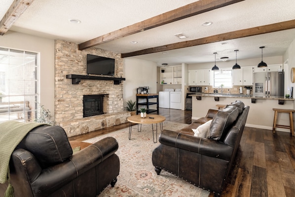 A living room with a stone fireplace, leather sofas, and a kitchen in the background. - The Getaway - Sleeps 12 (Belton)