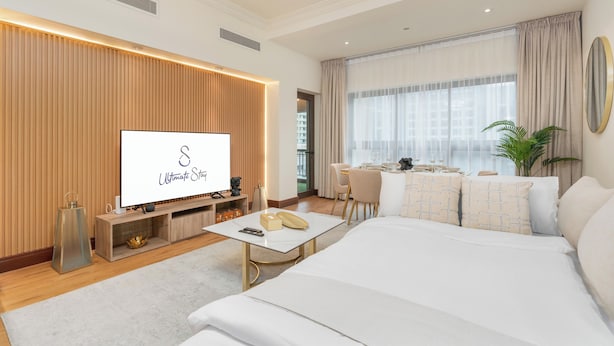 A modern hotel room with a large bed, a TV, a dining area, and a view of the city.