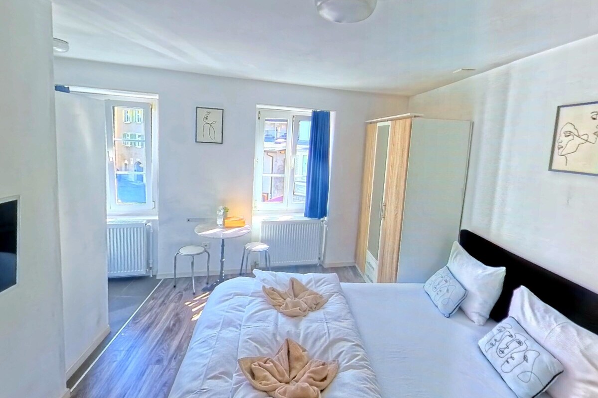 A hotel room with a large bed, a desk, two chairs, a radiator, and a window with curtains.